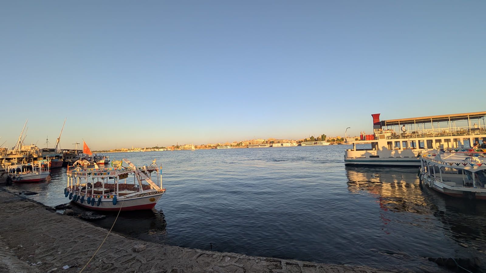 View across the Nile from Luxor’s West Bank beneath a wide blue sky, with boats on the river and the East Bank on the opposite shore.